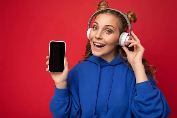 Piękny szczęśliwy uśmiech młoda kobieta noszenie stylowe casual strój odizolowany na tle ściany gospodarstwa i pokazując telefon komórkowy z pustym wyświetlaczem do makiety noszenia białe słuchawki bluetooth — Zdjęcie stockowe