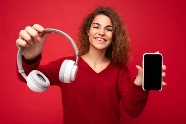 붉은 색 바탕에 고립된 짙은 빨간색 스웨터를 입은 젊은 여성이 웃으며 흰 무선 헤드폰 과빈 화면을 가진 휴대 전화를 제안 했다. — 스톡 사진