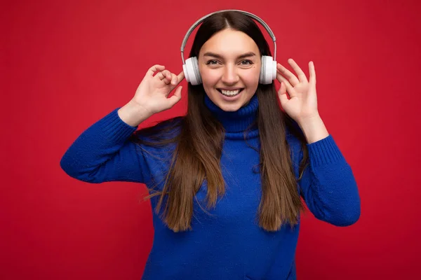 Attractive positive smiling young brunette woman wearing blue sweater isolated over red background wall wearing white bluetooth earphones listening to cool music and having fun looking at camera