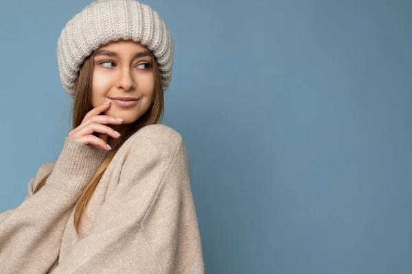 Portrait shot of beautiful sexy smiling happy young blonde woman standing isolated on blue background wall wearing beige sweater and beige hat looking to the side and flirting — Stock Photo, Image