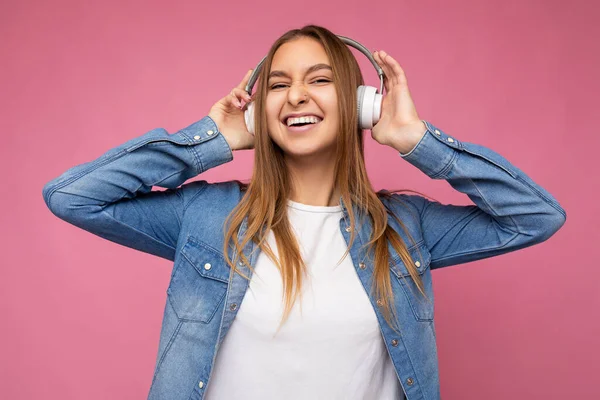 Photo of beautiful happy smiling young blonde woman wearing blue jean shirt and white t-shirt isolated over pink background wearing white wireless bluetooth earphones listening to cool music and — Stock Photo, Image