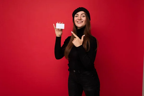 Fotografie krásné mladé brunetky žena na sobě černý svetr a klobouk izolované na červeném pozadí držící kreditní kartu při pohledu na kameru a ukazující prstem na plastovou kartu — Stock fotografie