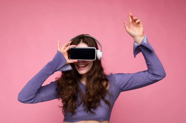 Güzel, mutlu, esmer, kıvırcık saçlı bir kadının pembe arka plan duvarında izole edilmiş bir şekilde çekilmiş fotoğrafı. Boş ekran ve bluetooth kulaklıklı cep telefonunu işaret ediyor.