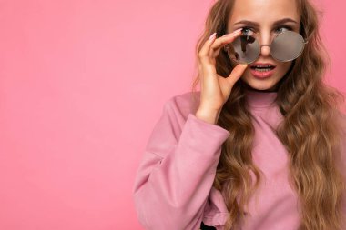 Yakından çekilen fotoğrafta güzel, koyu renk bir sarışın kadın günlük kıyafetler giyiyor ve renkli arka planda izole edilmiş güneş gözlüğü takıyor.