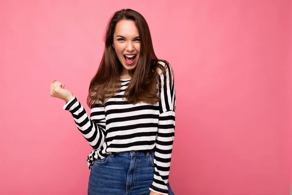 Portrait de jeune émotionnel positif heureux belle femme brune mignonne avec des émotions sincères dans pull rayé décontracté isolé sur fond rose avec de l'espace libre et de célébrer la victoire en criant oui — Photo