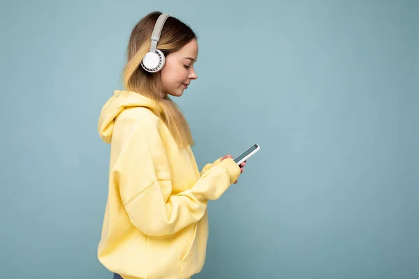 Foto de archivo lateral de una hermosa mujer joven que usa un elegante atuendo casual aislado sobre una colorida pared de fondo que usa auriculares inalámbricos blancos y escucha música y usa teléfono móvil — Foto de Stock