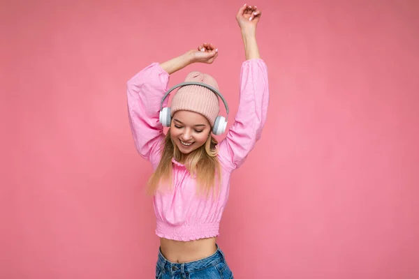 Beautiful happy smiling young blonde woman wearing pink blouse and pink hat isolated over pink background wall wearing white wireless bluetooth headphones listening to cool music having fun and — Stock Photo, Image