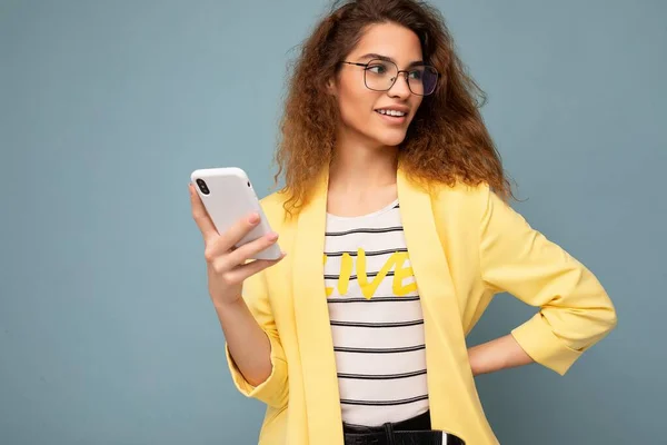 Portrait of attractive young woman with curly dark blond hair wearing yellow jacket and optical glasses isolated on background holding and using phone looking to the side — Stock Photo, Image