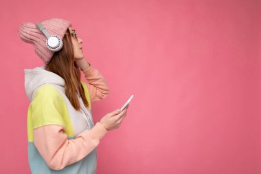 Yan profil fotoğrafı, arka plan duvarından izole edilmiş şık giyinen güzel, güler yüzlü genç bir kadın. Cep telefonu kullanıyor, beyaz bluetooth kulaklık takıyor.