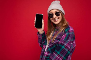 Güzel, gülümseyen, pozitif gülümseyen, mor gömlekli, sıradan beyaz tişörtlü, gri şapkalı ve güneş gözlüklü genç bir kadının fotoğrafı. Kırmızı arka planda izole edilmiş, el ele tutuşmuş ve cep telefonunu gösteriyor.