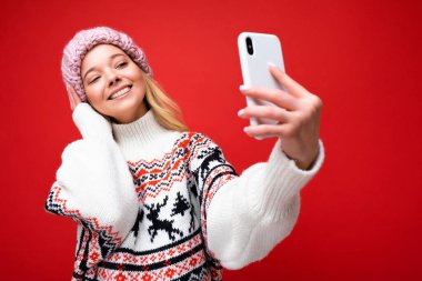 Oldukça olumlu sarışın bir kadının, kalın örgü şapkalı ve kalın kazaklı fotoğrafı. Kırmızı arka planda izole bir şekilde duruyor. Cep telefonuyla fotoğraf çekiyor.