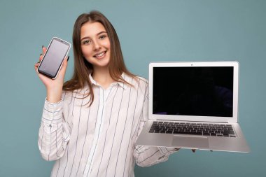 Mavi arka plan duvarında izole edilmiş, akıllı telefon ve bilgisayar laptopu tutan, cep telefonu ve netbook 'u boş gösteren, gündelik beyaz tişörtlü, çekici genç bir kadının fotoğrafı.