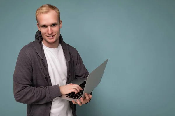 Boczny profil zdjęcie przystojny pewny siebie blondynka mężczyzna trzyma komputer laptop wpisując na klawiaturze w białym t-shirt i szary sweter patrząc na aparat odizolowany na niebieskim tle — Zdjęcie stockowe