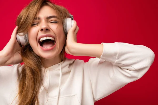 Shot of attractive crazy positive young blonde woman wearing white hoodie isolated over colourful background wearing white wireless bluetooth headsets listening to pop music and having fun — Stock Photo, Image