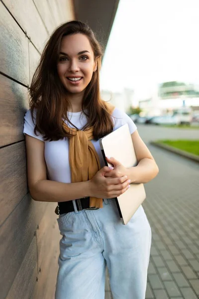 Krásná půvabná půvabná půvabná okouzlující veselá veselá rovnovlasá brunetka dívající se na kameru na ulici s notebookem v rukou s bílým tričkem a modrou — Stock fotografie