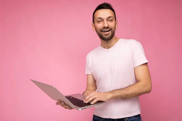 Guapo sonriente morena hombre sosteniendo ordenador portátil mirando a la cámara en camiseta sobre fondo rosa aislado — Foto de Stock