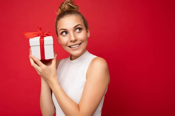 Piękne szczęśliwy młoda blondynka kobieta izolowane nad kolorowe tło ściany noszenie stylowe ubrania casual trzymając pudełko prezentów i patrząc na bok — Zdjęcie stockowe