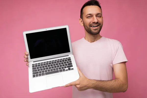 Bonito homem segurando computador portátil olhando para a câmera em t-shirt no fundo rosa isolado — Fotografia de Stock