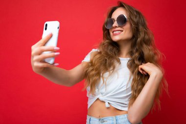 Çekici, genç, gülümseyen, mutlu bir kadın cep telefonu tutuyor ve kullanıyor. Selfie çekiyor. Duvar arkasından izole edilmiş şık kıyafetler giyiyor.