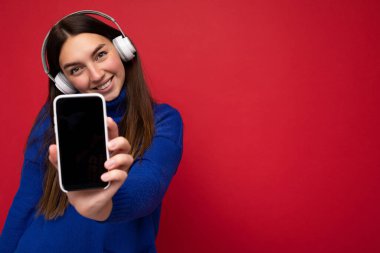 Güzel, mutlu, güler yüzlü, şık kıyafetler giyen, arka plandaki duvarda izole edilmiş, cep telefonlu beyaz kulaklık takan, model için boş bir ekran gösteren genç bir kadın.