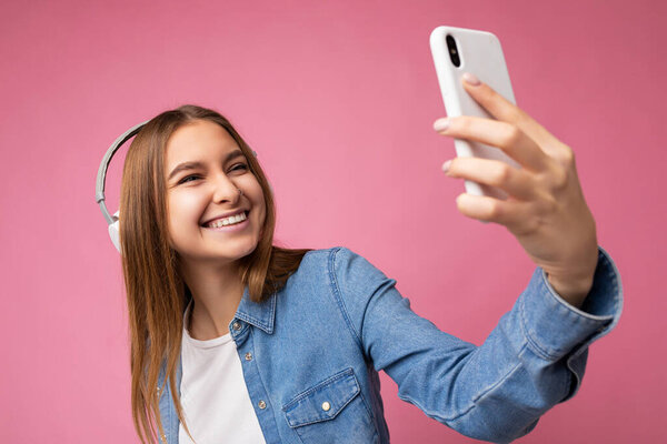 Photo of beautiful happy smiling young blonde woman wearing blue jean shirt and white t-shirt isolated over pink background wearing white bluetooth headphones and listening to music and using mobile