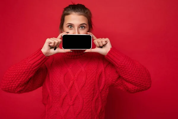 빨간 스웨터를 입고 빨간 배경 벽에 고립된 아름다운 젊은 여성의 사진 스마트폰을 들고 있고 카메라를 보기 위해 빈 화면을 보여 주는 핸드폰 — 스톡 사진