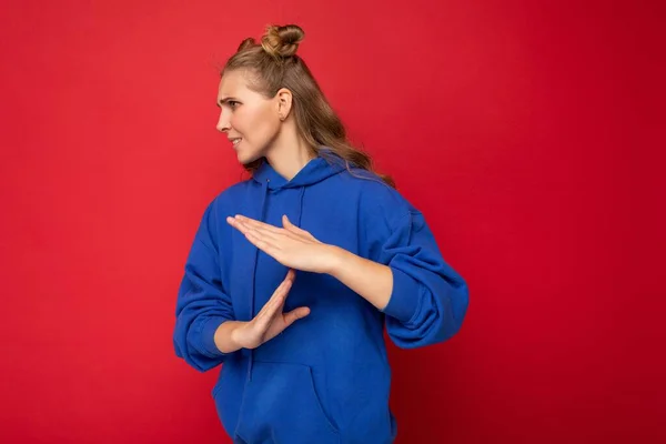Photo of emotional young attractive blonde woman with two horns with sincere emotions wearing hipster bright blue hoodie isolated over red background with copy space and showing pause sign — Stock Photo, Image