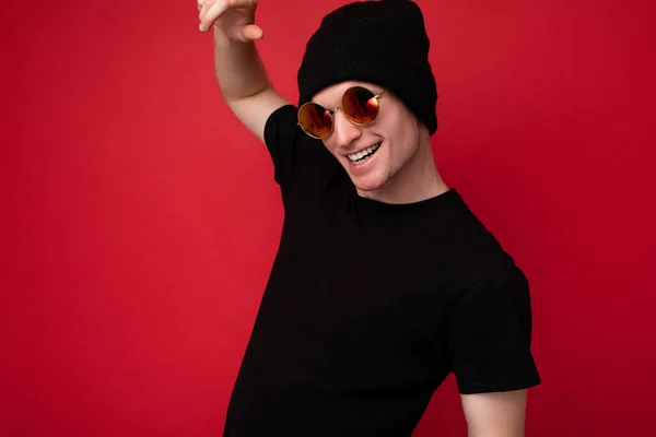 Photo shot of handsome smiling young man wearing black t-shirt for mockup black hat and stylish sunglasses isolated over red background wall looking at camera and having fun