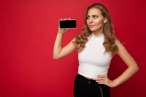 Mooie jonge blonde vrouw dragen witte t-shirt geïsoleerd op rode achtergrond met kopieerruimte houden smartphone tonen telefoon in de hand met leeg scherm voor mockup kijken naar gadjet display — Stockfoto