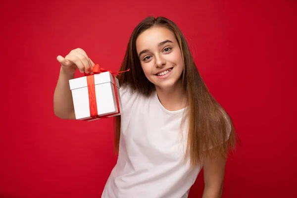 Vacker glad brunett flicka isolerad över färgglada bakgrund vägg bär eleganta casual kläder innehav presentförpackning och tittar på kameran — Stockfoto