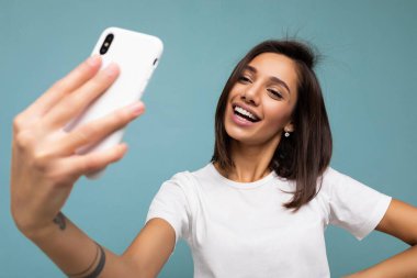 Çekici, genç, gülümseyen, mutlu bir kadın cep telefonu tutuyor ve kullanıyor. Selfie çekiyor. Duvar arkasından izole edilmiş şık kıyafetler giyiyor.