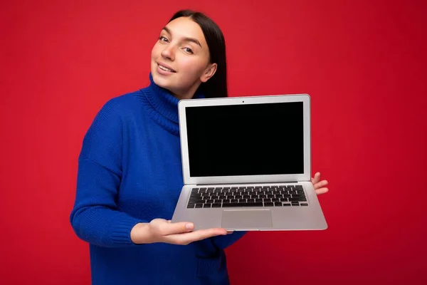 Encantador bastante feliz hermosa joven morena estudiante mujer sosteniendo ordenador portátil mirando a la cámara en suéter azul aislado sobre fondo de pared roja — Foto de Stock