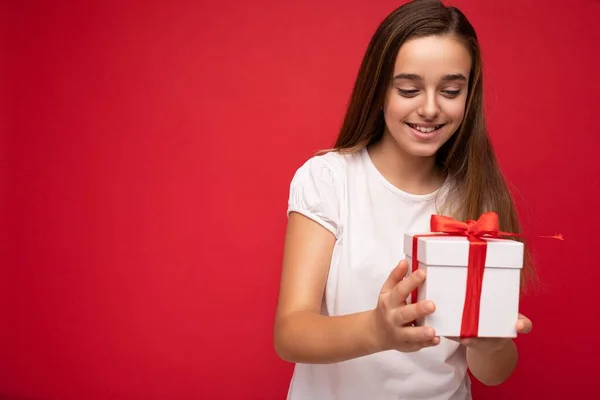 Foto de un hermoso niño femenino feliz positivo aislado sobre un bacground rojo que usa una camiseta blanca casual, una caja de regalo y se divierte. —  Fotos de Stock