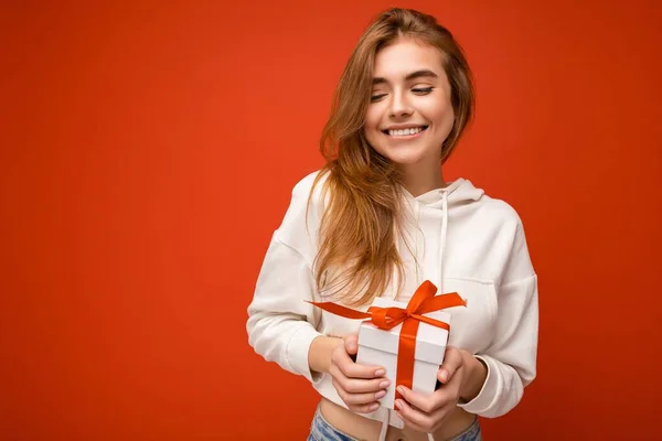 Fénykép lövés bájos boldog vidám mosolygós szőke fiatal nő elszigetelt át színes háttér fal visel fehér kapucnis gazdaság fehér ajándék doboz piros szalaggal, és szórakozás — Stock Fotó