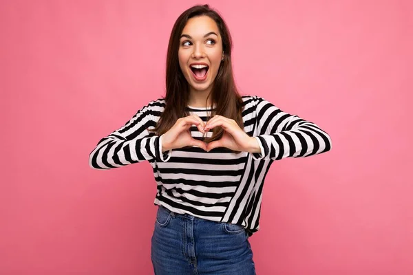 Young positive delightful happy nice cute brunette woman with sincere emotions wearing casual striped pullover isolated on pink background with empty space and making heart form with hands — Stock Photo, Image