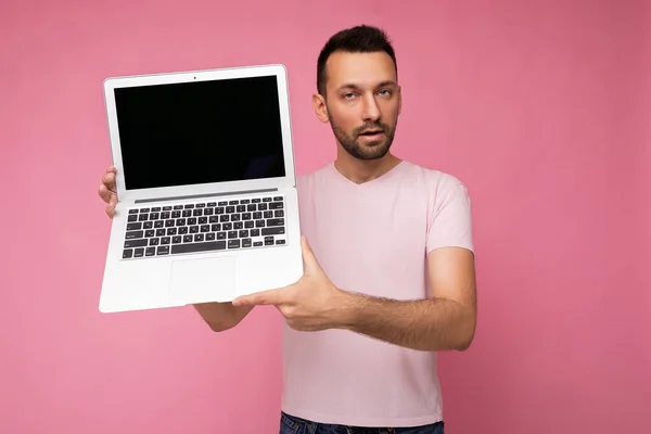 Hombre morena guapo sosteniendo ordenador portátil mirando a la cámara en camiseta sobre fondo rosa aislado —  Fotos de Stock