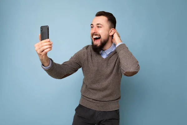 Feliz sonriente joven guapo usando ropa elegante casual de pie aislado sobre la pared de fondo sosteniendo el teléfono inteligente tomando foto selfie mirando la pantalla del teléfono móvil —  Fotos de Stock