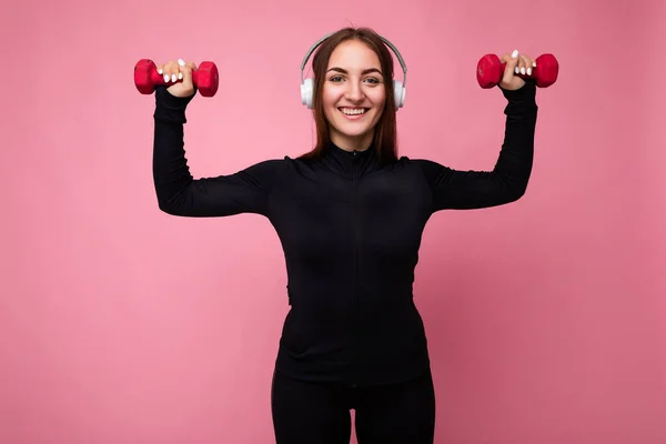 Krásná pozitivní úsměv mladá bruneta žena na sobě černé sportovní oblečení izolované přes růžové pozadí zeď dělá fitness pomocí činky na sobě bílé bluetooth sluchátka poslech dobré hudby — Stock fotografie
