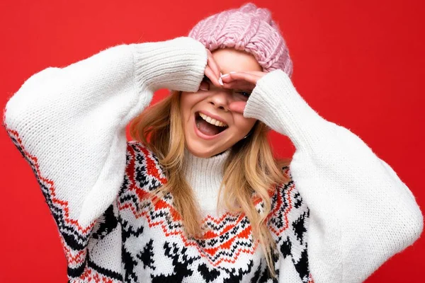 Primer plano de joven positiva hermosa mujer rubia con emociones sinceras usando sombrero de punto rosa y jersey de invierno aislado sobre fondo rojo y mostrando gafas con las manos —  Fotos de Stock