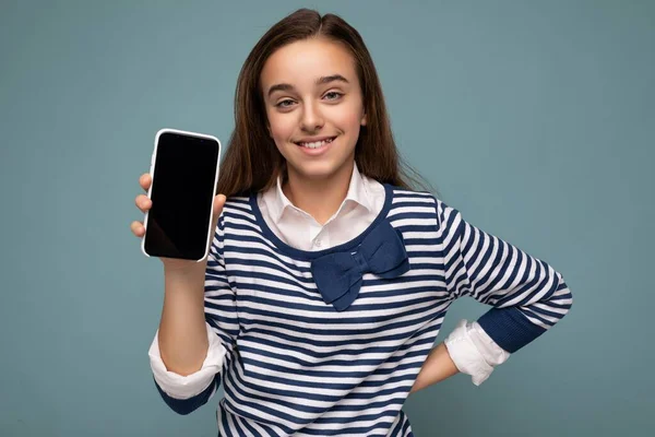 Chica sonriente bastante positiva con manga larga a rayas de pie aislada sobre fondo azul con espacio para copiar el teléfono inteligente que muestra el teléfono en la mano con pantalla vacía para maqueta apuntando a —  Fotos de Stock