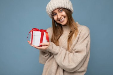 Çekici, mutlu, gülümseyen, mavi arka plan duvarında izole edilmiş, bej kazak ve bej örülmüş şapka giyen, elinde kırmızı kurdeleli beyaz bir hediye kutusu olan genç bir kadının fotoğrafı.