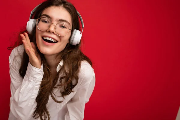 Closeup photo of attractive positive smiling young brunet woman wearing white shirt and optical glasses isolated over red background wearing white wireless bluetooth headsets listening to music and — Stock Photo, Image