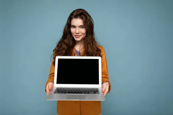 Foto de Hermosa mujer joven feliz seria sosteniendo computadora portátil mirando a la cámara con chaqueta amarilla aislada sobre fondo de pared azul —  Fotos de Stock