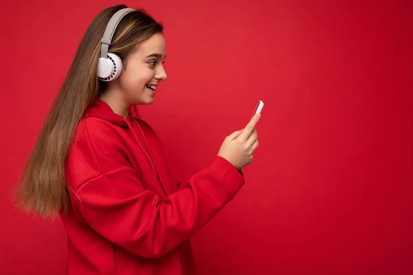Boczny profil Strzał pozytywnej uśmiechniętej ładnej brunetki w czerwonej bluzie odizolowanej na czerwonym tle, trzymającej wiadomość SMS w smartfonie z białymi słuchawkami bezprzewodowymi — Zdjęcie stockowe