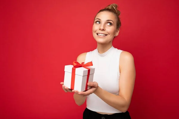 Bájos pozitív mosolygós fiatal szőke nő elszigetelt felett piros háttér fal visel fehér felső gazdaság ajándék doboz és felnéz, és álmodik — Stock Fotó