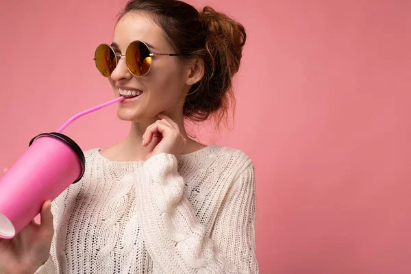 Primer plano Foto de atractiva joven feliz morena sonriente con suéter blanco y gafas de sol de colores aislados sobre fondo rosa sosteniendo taza de papel de café para cortar beber y — Foto de Stock