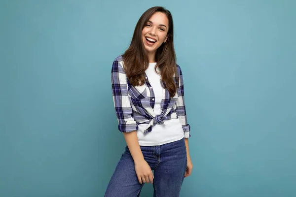 Portrait photo de jeune belle femme brune hipster souriante en chemise et jeans bleu et blanc tendance. Sexy insouciante femme posant isolé près du mur bleu avec espace vide en studio — Photo