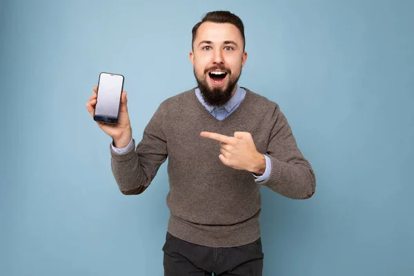 Amazed shocked Handsome happy cool young brunette unshaven man with beardwearing stylish grey sweater and blue shirt standing isolated over pink background wall holding smartphone and showing phone — Stock Photo, Image