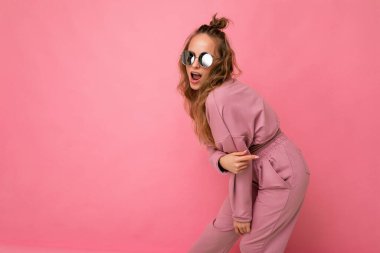 Güzel, mutlu, esmer, kıvırcık saçlı bir kadının pembe arka plan duvarında izole edilmiş, günlük pembe spor kıyafetleri ve şık güneş gözlükleriyle kameraya bakıp dans ettiği bir fotoğraf.