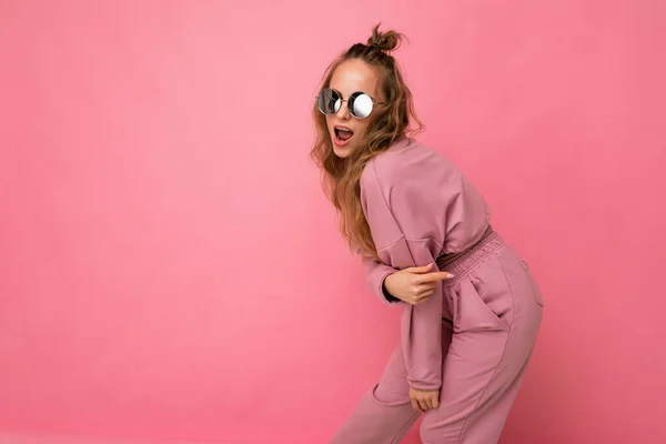 Skott av vackra glada unga mörka blonda lockigt kvinna isolerad över rosa bakgrund vägg bär casual rosa sport kläder och snygga solglasögon tittar på kamera och dans — Stockfoto